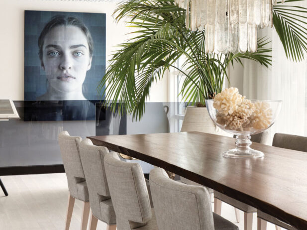 Room with long dark wood table, white chairs and a chandelier overtop with a palm plant and large black and white portrait in background