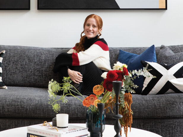 The designer sitting on a grey couch with vases of flowers and magazines on a coffee table in front.